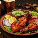 Prato indiano com frango Tandoori assado servido com rodelas de limão, cebola roxa, molho chutney verde e especiarias, acompanhado por lassi em canecas de cobre.
