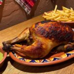 Pollo a la Brasa dourado com pele crocante servido em prato de cerâmica decorado com padrões geométricos, acompanhado de batatas fritas douradas, molho aji verde em tigela de barro e salada fresca, sobre mesa de madeira rústica com têxtil peruano tradicional ao fundo