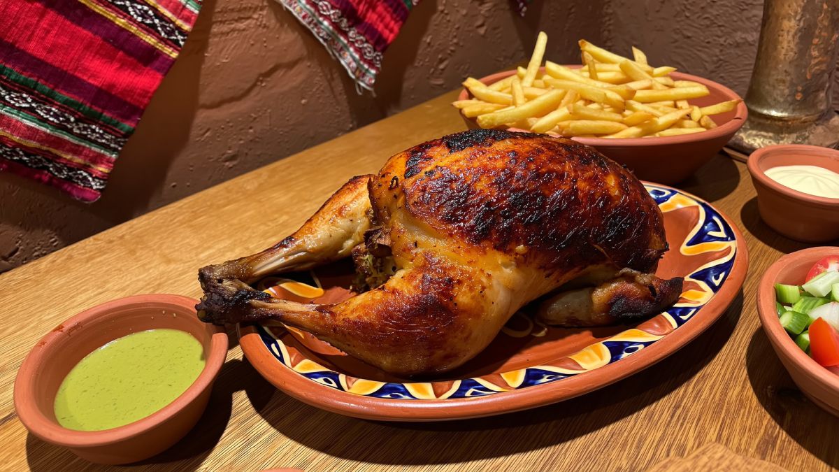Pollo a la Brasa dourado com pele crocante servido em prato de cerâmica decorado com padrões geométricos, acompanhado de batatas fritas douradas, molho aji verde em tigela de barro e salada fresca, sobre mesa de madeira rústica com têxtil peruano tradicional ao fundo