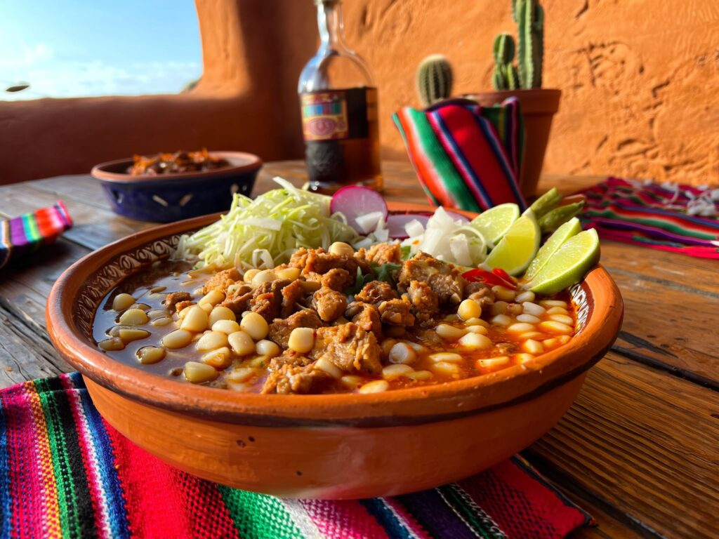 Prato de Pozole Mexicano servido em tigela de cerâmica com milho branco, carne, repolho, limão, cebola e pimenta, sobre mesa rústica com toalha colorida típica do México.