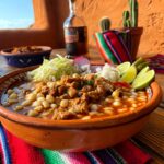 Prato de Pozole Mexicano servido em tigela de cerâmica com milho branco, carne, repolho, limão, cebola e pimenta, sobre mesa rústica com toalha colorida típica do México.
