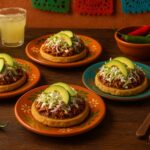 Quatro sopes mexicanos servidos em pratos coloridos, cobertos com carne desfiada, alface, cebola roxa, queijo branco e fatias de abacate, acompanhados por bebidas e pimentas sobre uma mesa rústica.