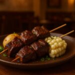 Anticuchos peruanos servidos em prato rústico de cerâmica, com espetos de carne grelhada, milho branco peruano e batata cozida, decorados com ervas frescas, sobre mesa de madeira em ambiente de restaurante aconchegante.
