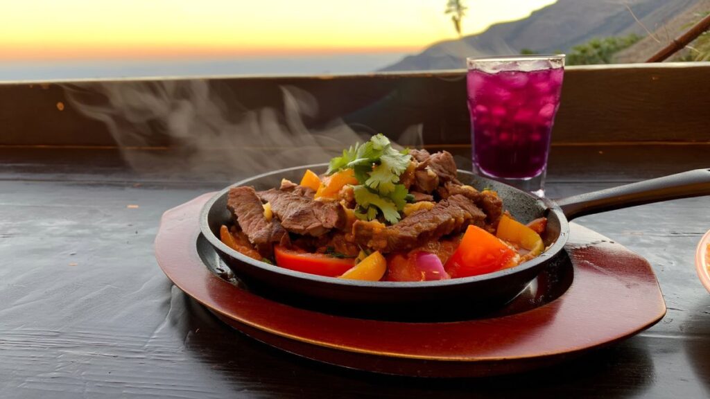 Prato típico de Lomo Saltado peruano servido em frigideira com carne salteada, pimentões coloridos, cebola roxa e coentro, acompanhado de bebida roxa com gelo ao fundo, em cenário ao pôr do sol.