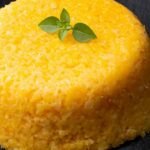 Cuscuz nordestino amarelo-dourado moldado em cilindro sobre base de ardósia preta, com brilho de manteiga na superfície e um pequeno ramo de folhas verdes como guarnição — textura granular visível.