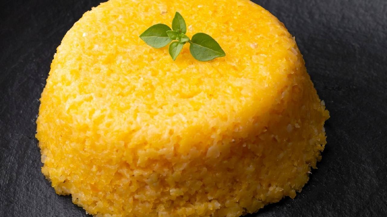 Cuscuz nordestino amarelo-dourado moldado em cilindro sobre base de ardósia preta, com brilho de manteiga na superfície e um pequeno ramo de folhas verdes como guarnição — textura granular visível.